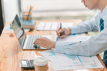 A man is sitting at a desk with a laptop and a notebook. He is writing on the notebook with a pen. The scene suggests that he is working on a project or a report, possibly related to his job