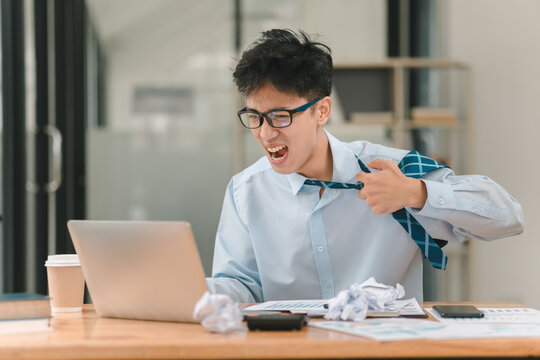 Frustration at the Deadline: A young businessman in a white shirt and tie, sits at his desk, throws his tie off in frustration, staring at his laptop. Papers are strewn across the desk, a crumpled bal