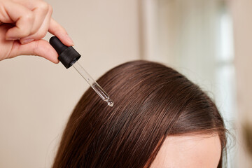 A young woman applies a drop of oil from a pipette to her hair, close-up. Vitamins, keratin for...