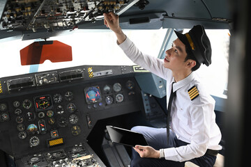 Pilot with digital tablet sitting in cockpit and adjusting controls on the overhead panel