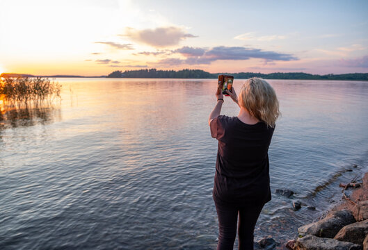 One caucasian woman on the lake shore admires the sunset and takes a selfie on a mobile phone.