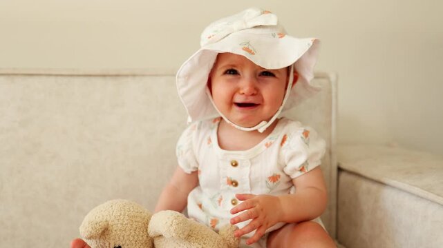 A little baby girl is sitting on the couch and crying. She wants to take off her sun hat and is crying hard. Baby tears.