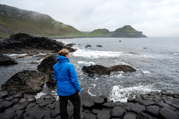 Frau mit blauer Regenjacke und blonden Haaren besichtigt die gleichmäßig geformten Basaltsäulen...