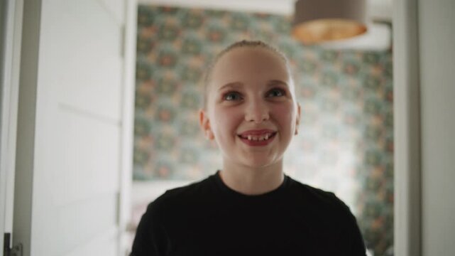 Adorable and happy young girl with make up and bun hairstyle makes funny hand gestures on camera. Cheerful Caucasian teenager smiles and looks at camera while having fun at home. Lifestyle concept.