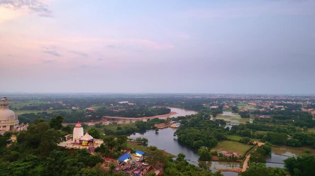 A drone shot of Dhauli hilltop shows two temples amid lush greenery under a pink orange dawn or dusk sky, with a winding river and distant fields adding to the serene atmosphere. Odisha