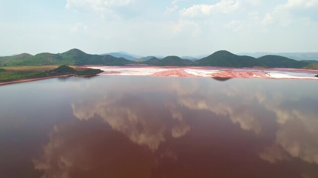 aerial view of red mud pond in Koraput, Odisha, calm, reflective water bordered by reddish-brown and white sediment, with green rolling hills in the background under a partly cloudy sky.