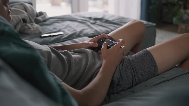 Close up of Caucasian teenager playing online video game on console using wireless controller, lying on sofa in comfort living room. Young boy relaxing and spending weekend at home. Lifestyle concept.