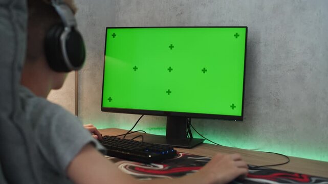 Caucasian teenager wearing headphones looks at personal computer monitor with green screen. Teen boy watches video or plays video game on PC spending time at home. Chroma key template screen. Mock up.