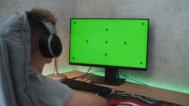 Caucasian teenager wearing headphones looks at personal computer monitor with green screen. Young boy watches video on PC spending time at home. Lifestyle concept. Chroma key template screen. Mock up.