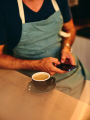 A barista in an apron uses a smartphone next to a freshly served cup of coffee, highlighting the blend of modern technology and traditional coffee service in a cafe setting.