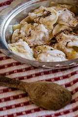 Traditional Circassian ravioli dish served in a metal bowl with a lid, accompanied by a glass of yogurt, with a wooden spoon and a red and white striped cloth.