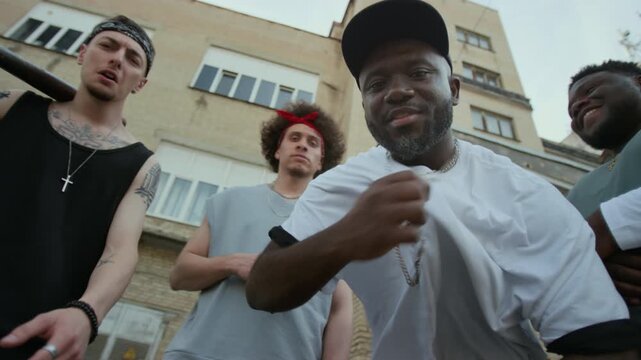 Young Black rapper wearing t-shirt, cross chain and cap performing on camera while standing in city street with hip hop band