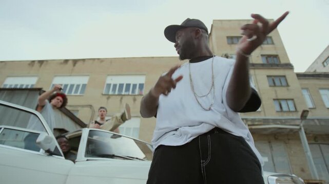 Low angle view of cool Black musician wearing t-shirt, cross chain and cap rapping on camera expressively while his hip hop group sitting in car behind in urban street