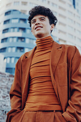 Young man in stylish brown outfit posing confidently in urban setting with modern buildings in the...