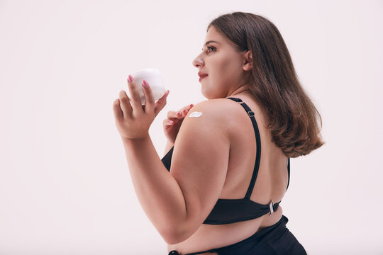 Rear view photo of chubby, young woman posing in black swimwear and applying cream to protect her skin from sun light against white studio background. Concept of natural beauty, body positivity, spa.
