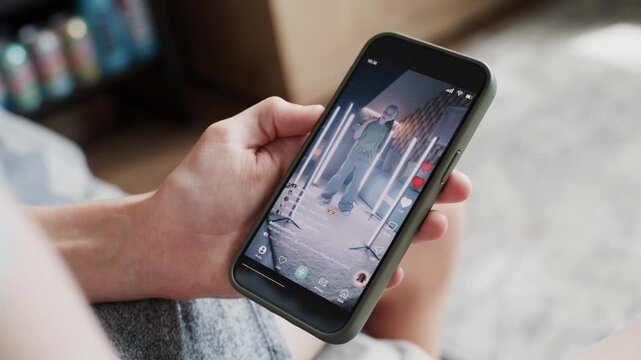 Social media feed with playback of dancing young girl video displayed on mobile phone screen. Teenage boy sits on sofa at home, surfs the Internet using smartphone, spends leisure time. Close Up.