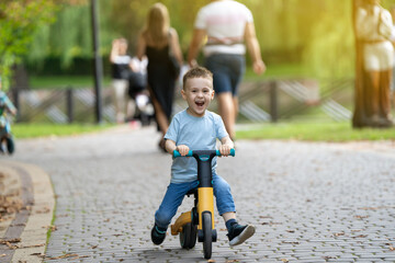 A cute happy caucasian toddler boy of three or four years old rides a bicycle or balance bike in the park against the backdrop of a crowd of people in the background. Summer activity for children.