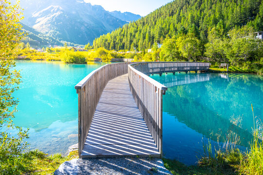 A tranquil wooden boardwalk bridge curves above the vibrant blue waters of Lake in Sulden, Italy. With the majestic Italian Alps towering in the background under a clear azure sky.