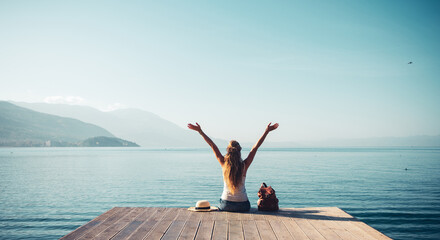 Woman with arms raised up sitting on wooden pier enjoying traveling, freedom, adventure in front of...