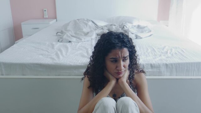 A wide shot of the young curly-haired pensive lonely anxious Indian Asian female or woman sitting on the floor in destress in the bedroom. Concept of heartbreak, depression, mental health medication