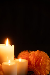 Vertical view of three lit candles on the left, surrounded by orange marigolds, all resting on a Day of the Dead-patterned tablecloth