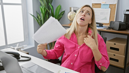 Young blonde woman in a pink shirt feeling hot in a cluttered office setting fanning herself with paper