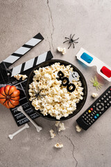 Vertical bowl of popcorn with Halloween decorations, a film clapperboard, 3D glasses, and a remote control for a horror movie night