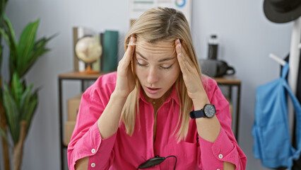 A stressed young blonde woman wearing a pink shirt feeling headache at her cluttered office space