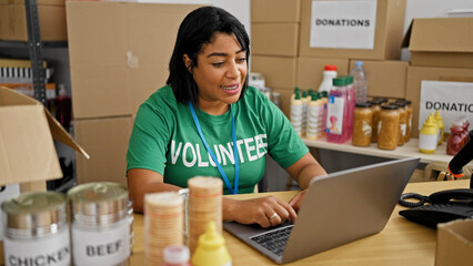 Hispanic woman volunteering at a donation center, organizing supplies and working on a laptop in a warehouse setting.