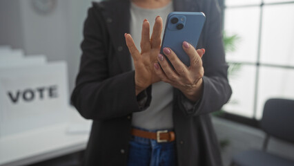 A mature woman checks her smartphone while standing in an indoor electoral college room, surrounded by voting booths.