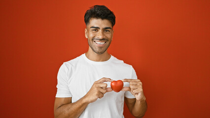 A smiling young hispanic man with a beard holds a heart shape isolated against a vibrant red...