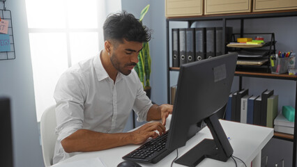 Handsome young man working in a modern office typing on a computer while seated at a desk with...