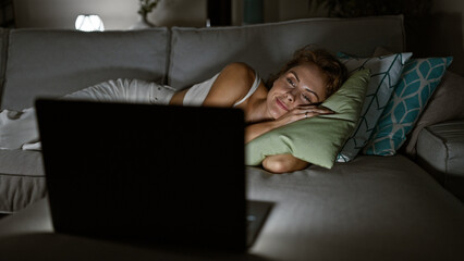 A young woman relaxes on a couch at home, watching a laptop in a cozy, dimly lit living room.