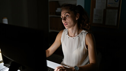 Focused caucasian woman working late in a dimly lit office on her computer, evoking dedication and...