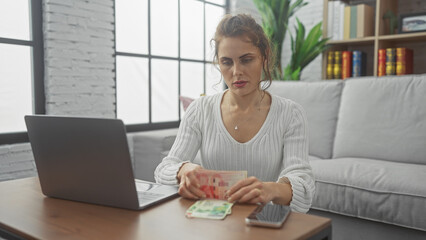 A focused young woman counts israeli shekels at home, seated by a laptop in a bright, cozy room.