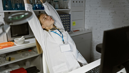 Fototapeta premium A tired, blonde woman scientist in a laboratory stretching with fatigue, surrounded by medical equipment.