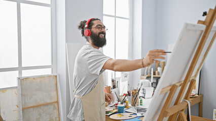 Hispanic man with beard painting in a bright art studio, wearing headphones and an apron.