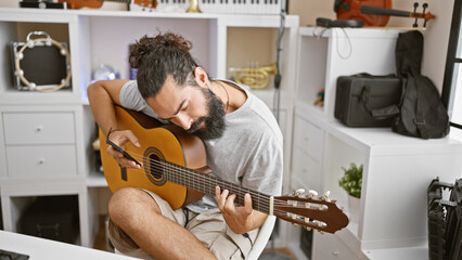 A bearded young man plays an acoustic guitar in a music-filled home studio setting.
