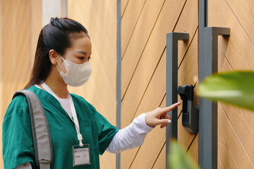 Asian woman wearing face mask using security access panel outdoors holding bag and ID badge while appearing focused on task at hand
