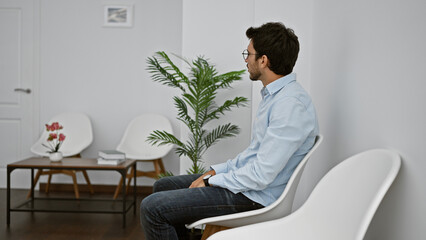 A bearded young man in a blue shirt sits contemplatively in a modernly furnished white room with plant decor.