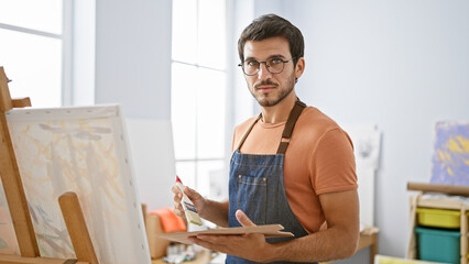 A young hispanic man with a beard and glasses painting on a canvas in an art studio, conveying creativity and concentration.