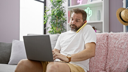 A middle-aged man works on laptop while holding a phone at shoulder in a cozy living room.