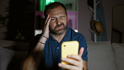 Mature man with grey hair feeling headache while looking at smartphone in dark living room.