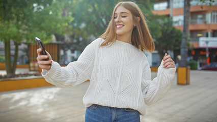 A joyful, young woman dances outdoors while holding a smartphone in a city park.