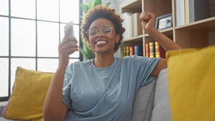 Joyful african woman with glasses celebrating success in a cozy living room.