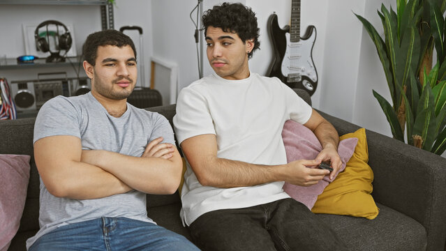 Two men lounging on a sofa in a modern living room with guitars and plants, portraying a relaxed atmosphere.