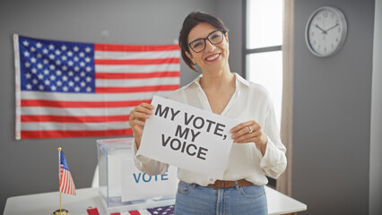 Hispanic woman holds a sign reading 'my vote, my voice' in a room with united states flags,...