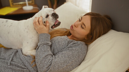 A young, beautiful, blonde woman lies in bed in her bedroom, gently playing with her canine pet, in a cozy home interior.