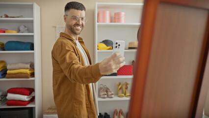 Handsome bearded hispanic man taking a selfie in wardrobe room with smartphone, wearing glasses and casual clothes.