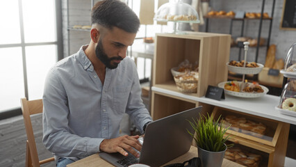 Young hispanic man with a beard working on a laptop in a cozy bakery filled with assorted pastries and natural light pouring in from large windows - Powered by Adobe